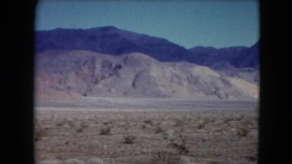 1961 Nevada 8mm frame showing scrubby desert foreground, pale foothills, and a darker mountain ridge beneath clear blue sky.
