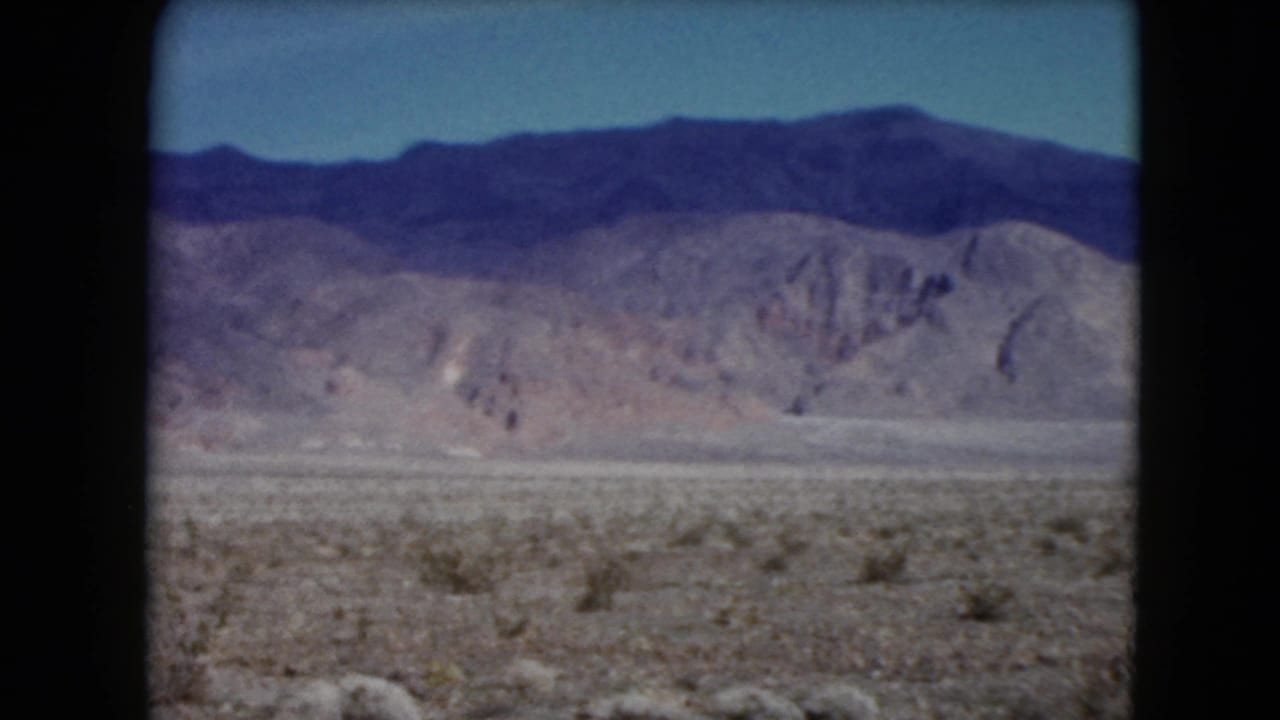 1961 Nevada home-movie frame with an open desert floor, distant foothills, and a broad dark ridge line under blue sky.
