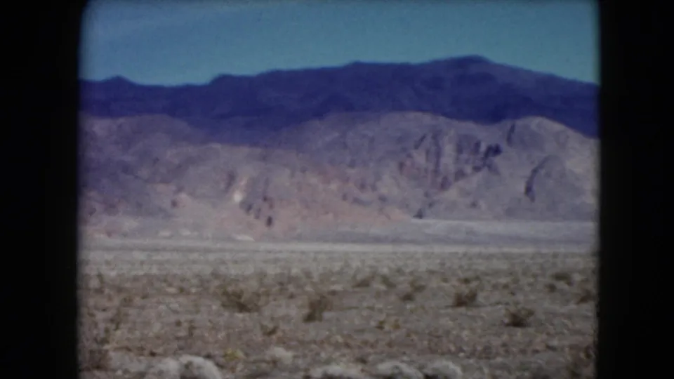 1961 Nevada home-movie frame with an open desert floor, distant foothills, and a broad dark ridge line under blue sky.