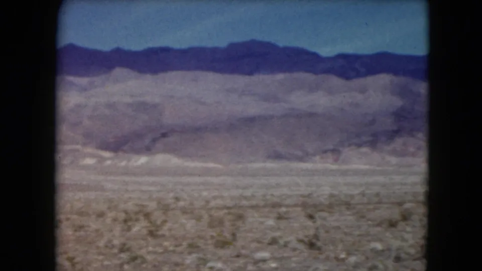 1961 Nevada 8mm frame showing a broad, empty desert basin with sparse shrubs and a long mountain range on the horizon.