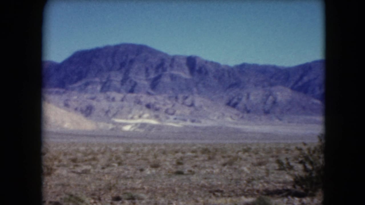 Open desert plain with low shrubs and rugged mountains under blue sky.