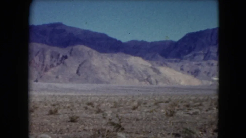 1961 Nevada 8mm frame showing scrubby desert ground, pale rocky hills, and darker mountains under a clear sky.