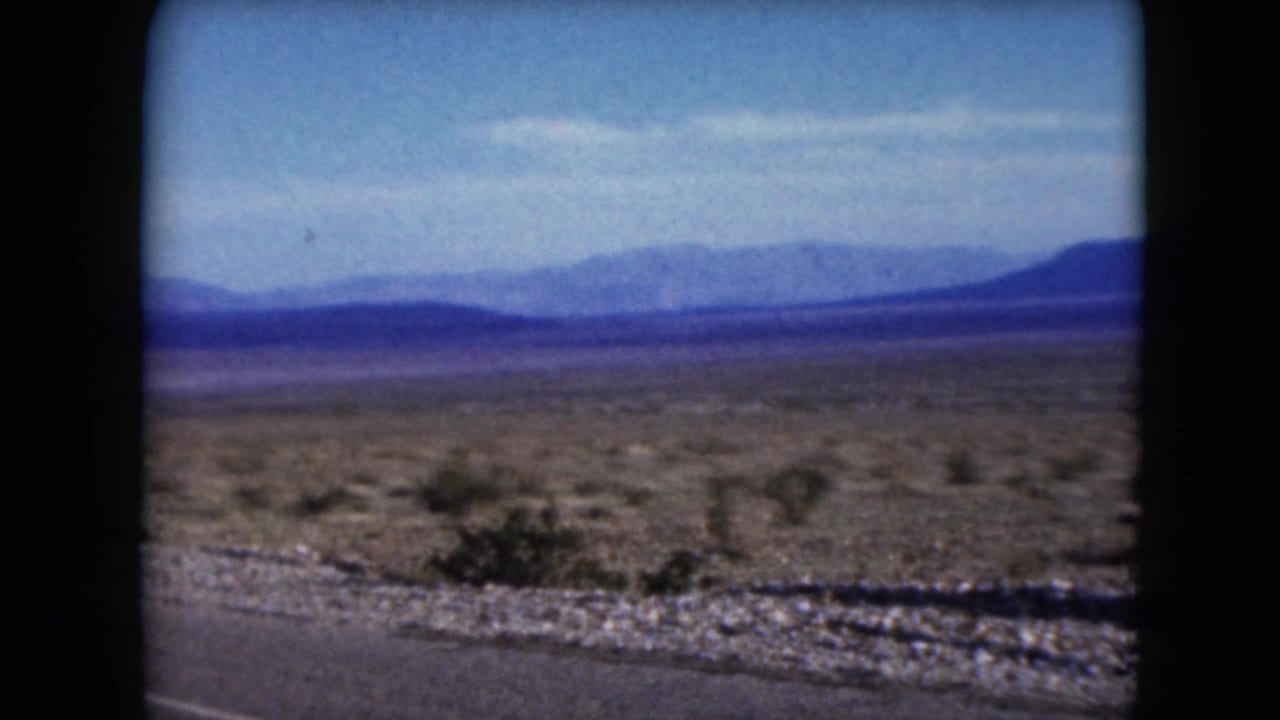 1961 Nevada 8mm frame showing a paved desert road edge, scrubland, and hazy mountain ridges in the distance.