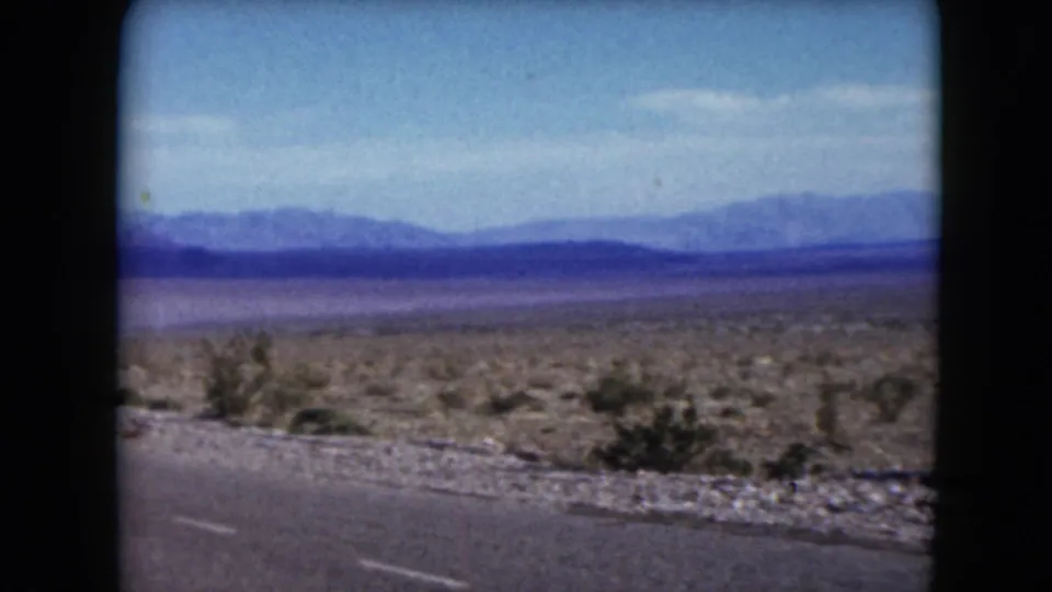 1961 Nevada travel footage frame with road shoulder, sparse desert brush, blue sky, and hazy mountains from a moving vehicle.