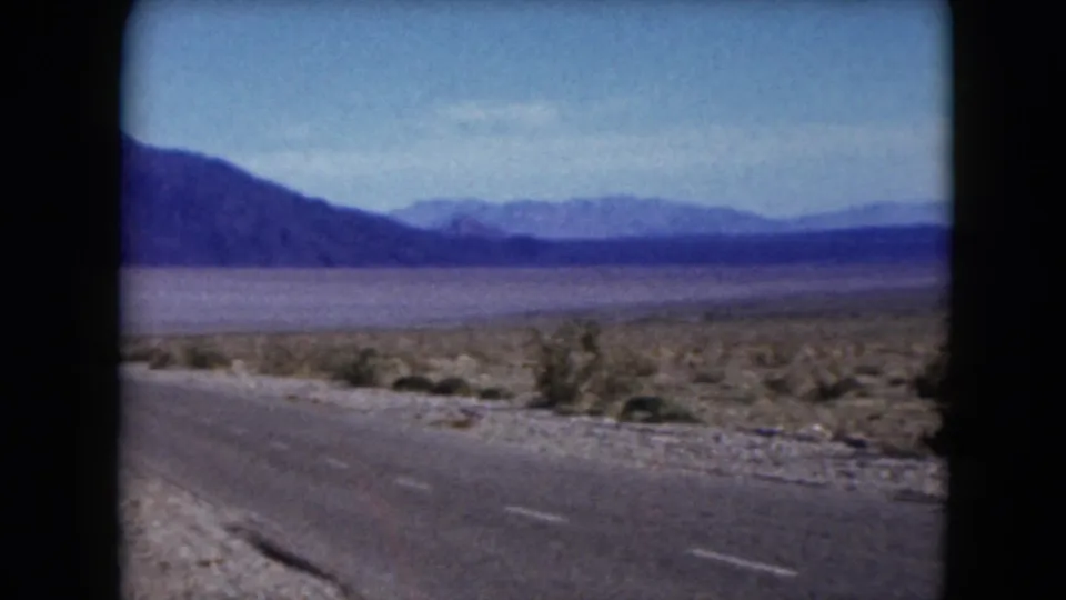 1961 Nevada 8mm frame showing a paved road, gravel shoulder, arid scrubland, and layered mountain ridges.