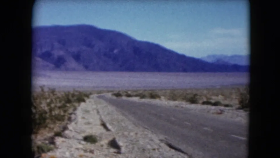 1961 Nevada 8mm frame showing a roadside view toward a prominent mountain ridge across dry desert terrain.
