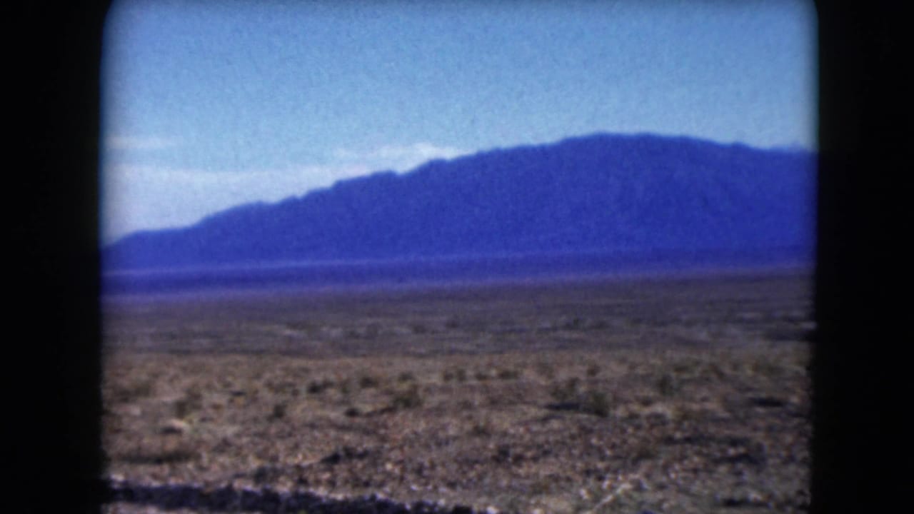 1961 Nevada 8mm frame showing open desert scrub, a low roadside foreground, and distant blue mountains beneath a clear pale sky.