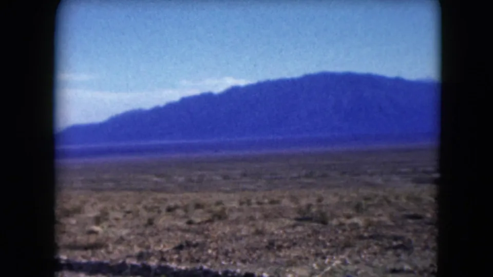 1961 Nevada 8mm frame showing open desert scrub, a low roadside foreground, and distant blue mountains beneath a clear pale sky.