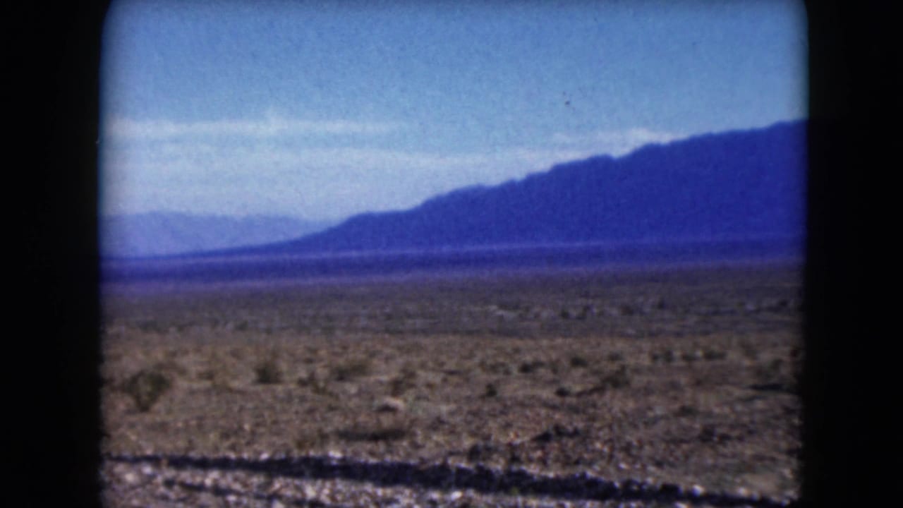 Wide desert plain with brush, roadside edge, and mountains stretching across the horizon.