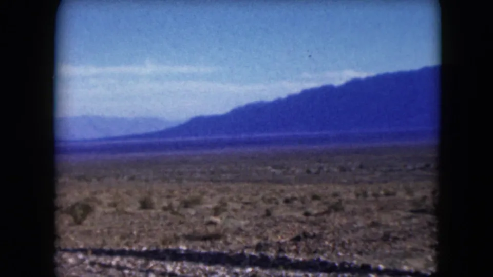 1961 Nevada 8mm frame from a moving vehicle, showing scrubby desert ground, a gravel roadside edge, and a dark mountain range in the distance.