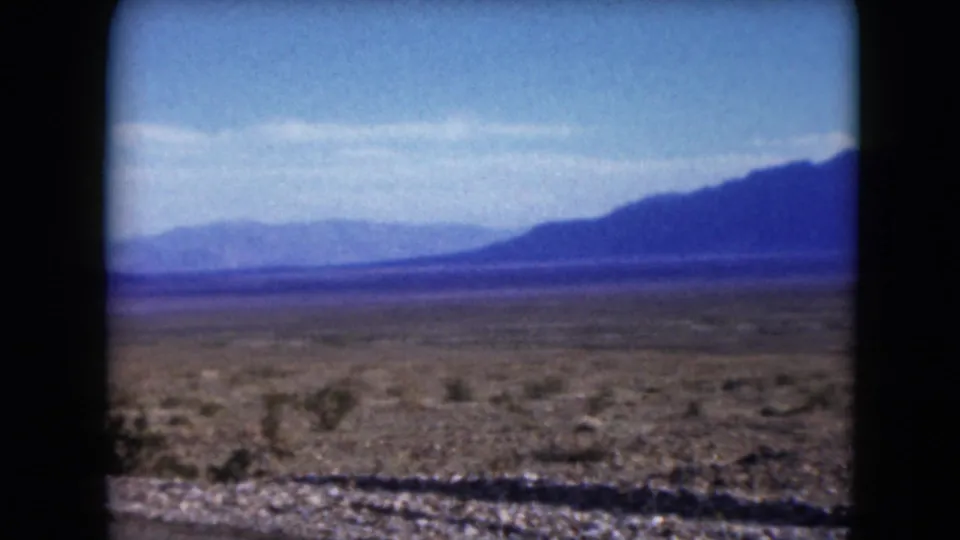 1961 Nevada 8mm frame showing a flat arid plain with scattered shrubs, roadside gravel, and hazy mountains beyond the horizon.