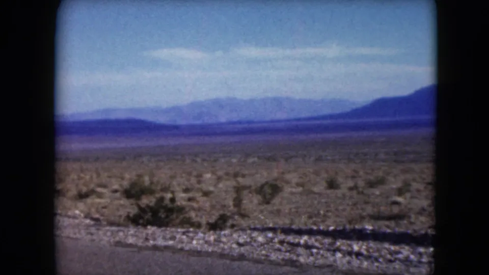 1961 Nevada 8mm frame showing open desert vegetation, a roadside strip, and layered blue mountains under a lightly hazy sky.