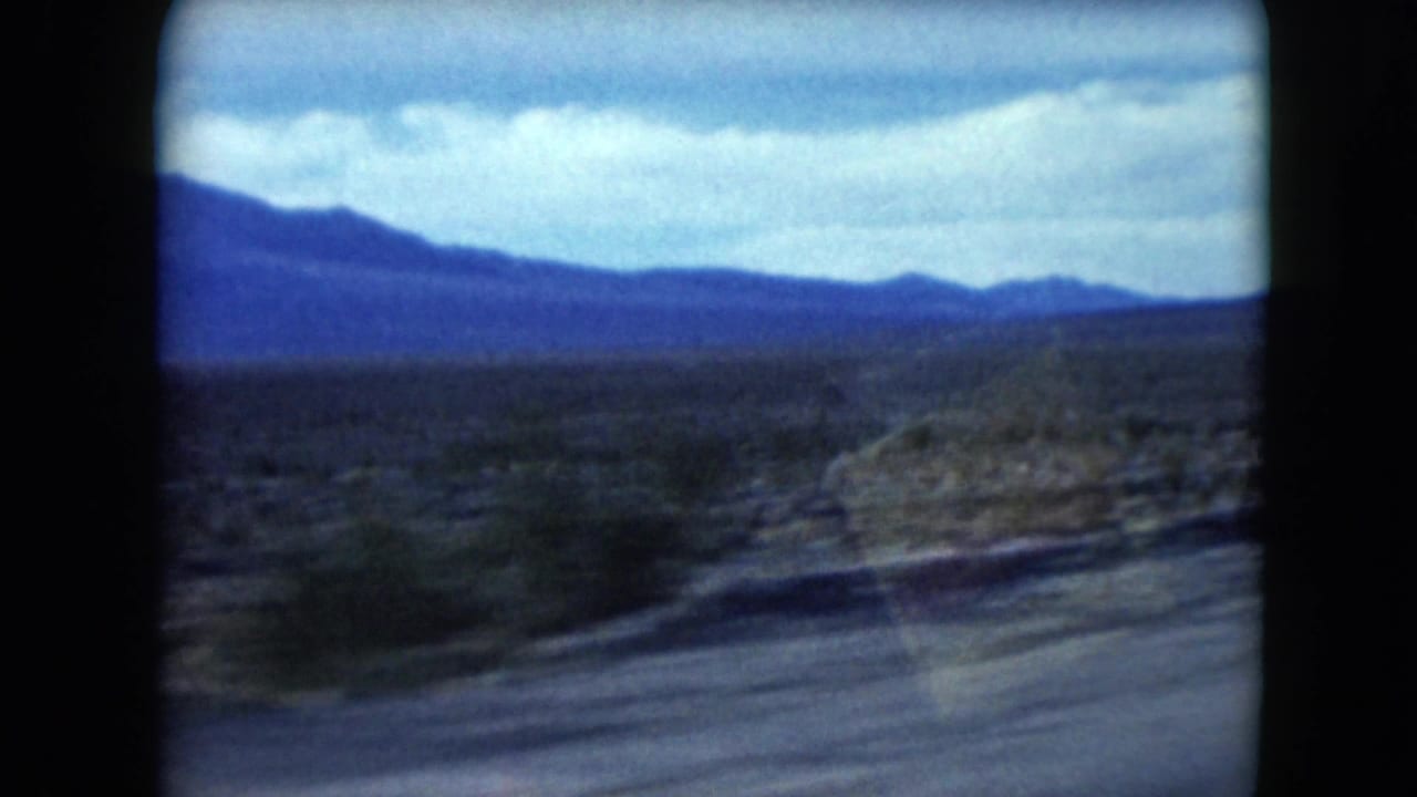 Desert roadside with low mountains under a cloudy sky, seen through 8mm frame edges.