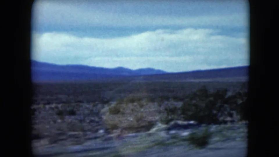 1961 Nevada desert road view with scrub, motion blur, and distant hills in 8mm home-movie color.
