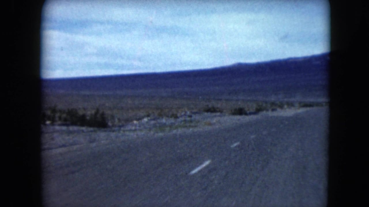Paved desert road leading toward low mountains under a pale sky.