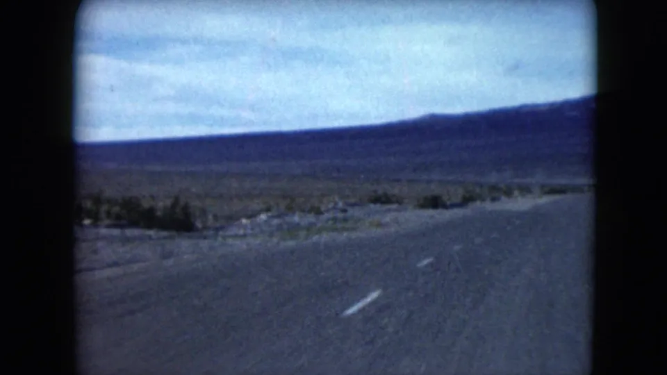 1961 Nevada 8mm frame showing a paved road crossing flat desert toward distant low mountains under a cloudy pale-blue sky.