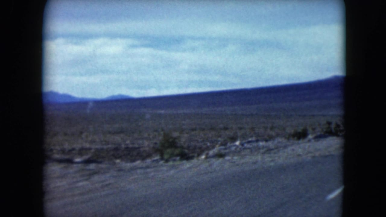 1961 Nevada road-trip frame with scrubby desert beside the pavement and distant hills softened by 8mm grain and motion.