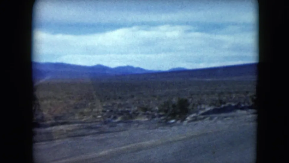 1961 Nevada 8mm frame showing open desert, a partial road edge, sparse shrubs, and blue distant mountain silhouettes.