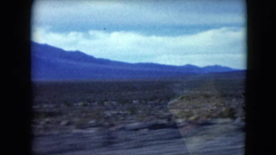 1961 Nevada desert view with a blue mountain range beyond scrubland and a blurred roadside foreground from vehicle motion.