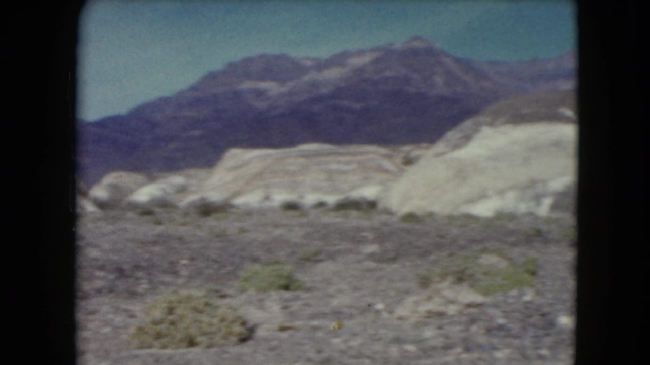Pale desert hills and distant mountains beyond gravelly ground and sparse shrubs.