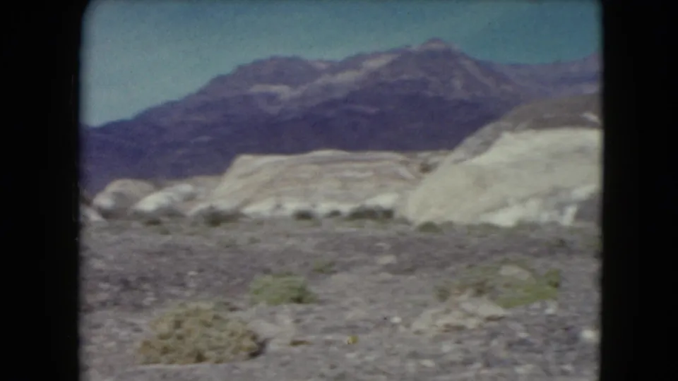 1961 Nevada 8mm frame showing pale desert hills, scrubby ground, and darker mountains under a blue sky.