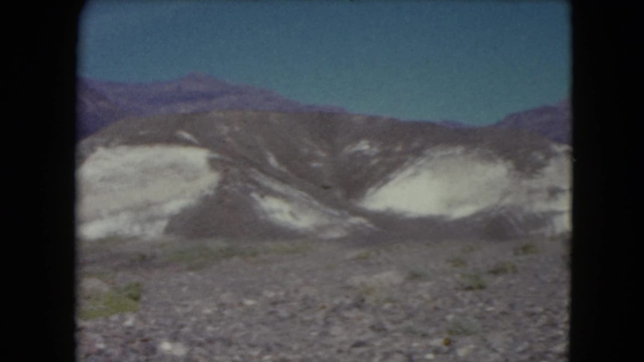 1961 Nevada travel frame with layered light rock formations, dark mountain slopes, and sparse desert vegetation.