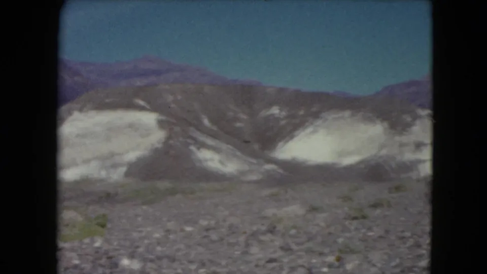 1961 Nevada travel frame with layered light rock formations, dark mountain slopes, and sparse desert vegetation.