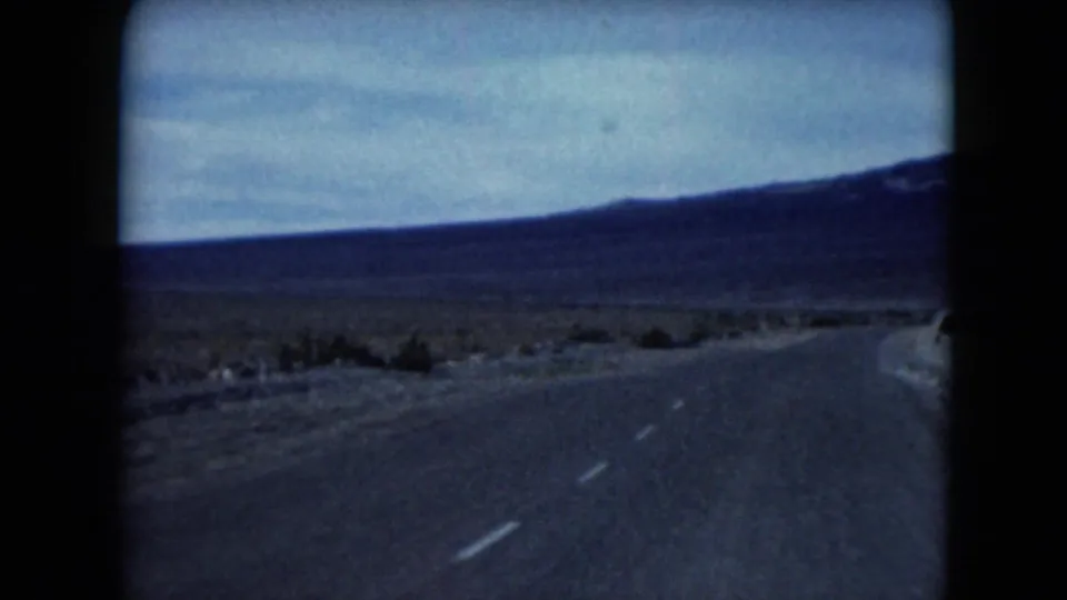 1961 Nevada home-movie frame looking down a paved desert road toward low mountains under a blue sky.