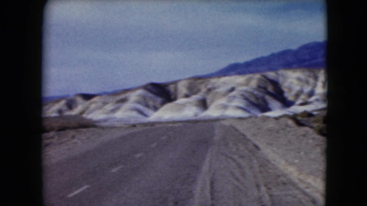 1961 Nevada home-movie frame showing a paved road and gravel shoulder approaching pale desert hills with mountains beyond.