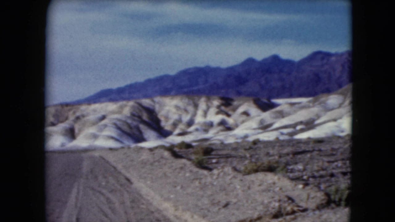 1961 Nevada frame with a road edge, rocky desert foreground, pale eroded hills, sparse shrubs, and a dark mountain ridge.
