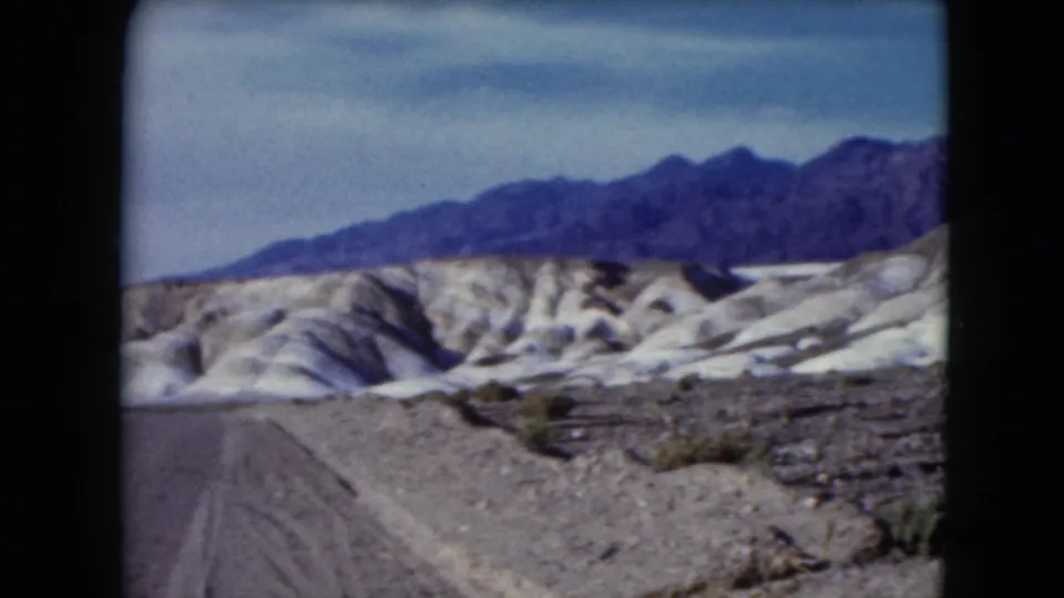 1961 Nevada frame with a road edge, rocky desert foreground, pale eroded hills, sparse shrubs, and a dark mountain ridge.