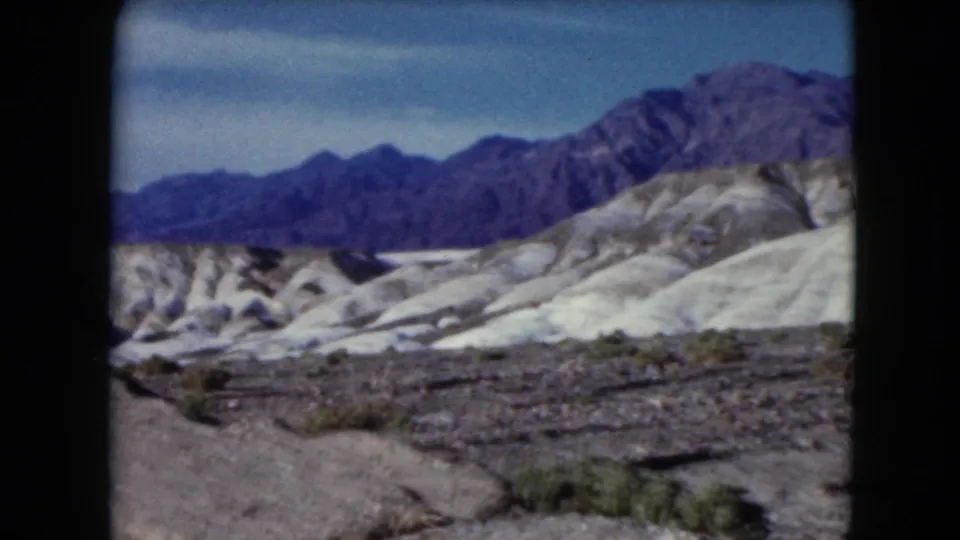 1961 Nevada 8mm view across rocky roadside terrain, sparse vegetation, pale hills, and blue-toned mountains.