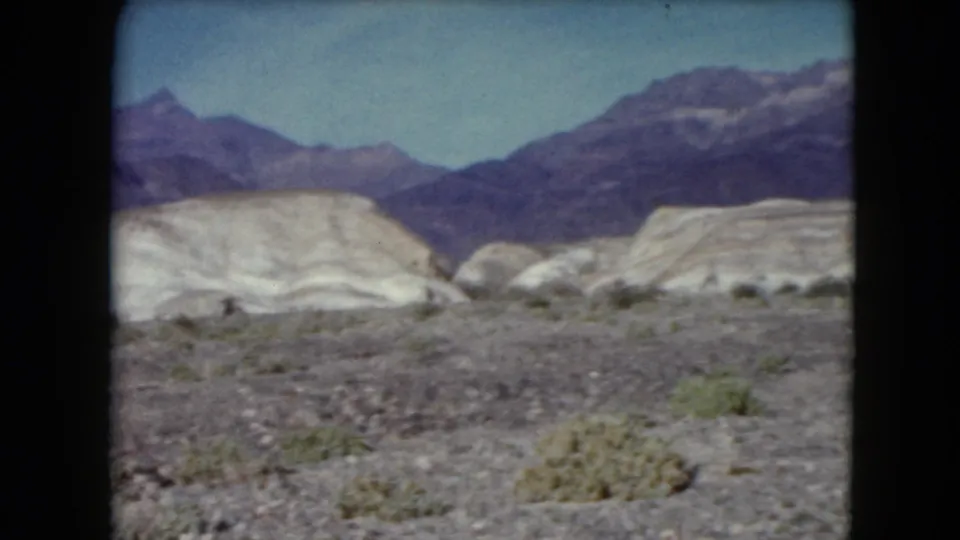 1961 Nevada landscape frame showing open rocky desert, scattered shrubs, pale ridges, and layered mountains in the distance.