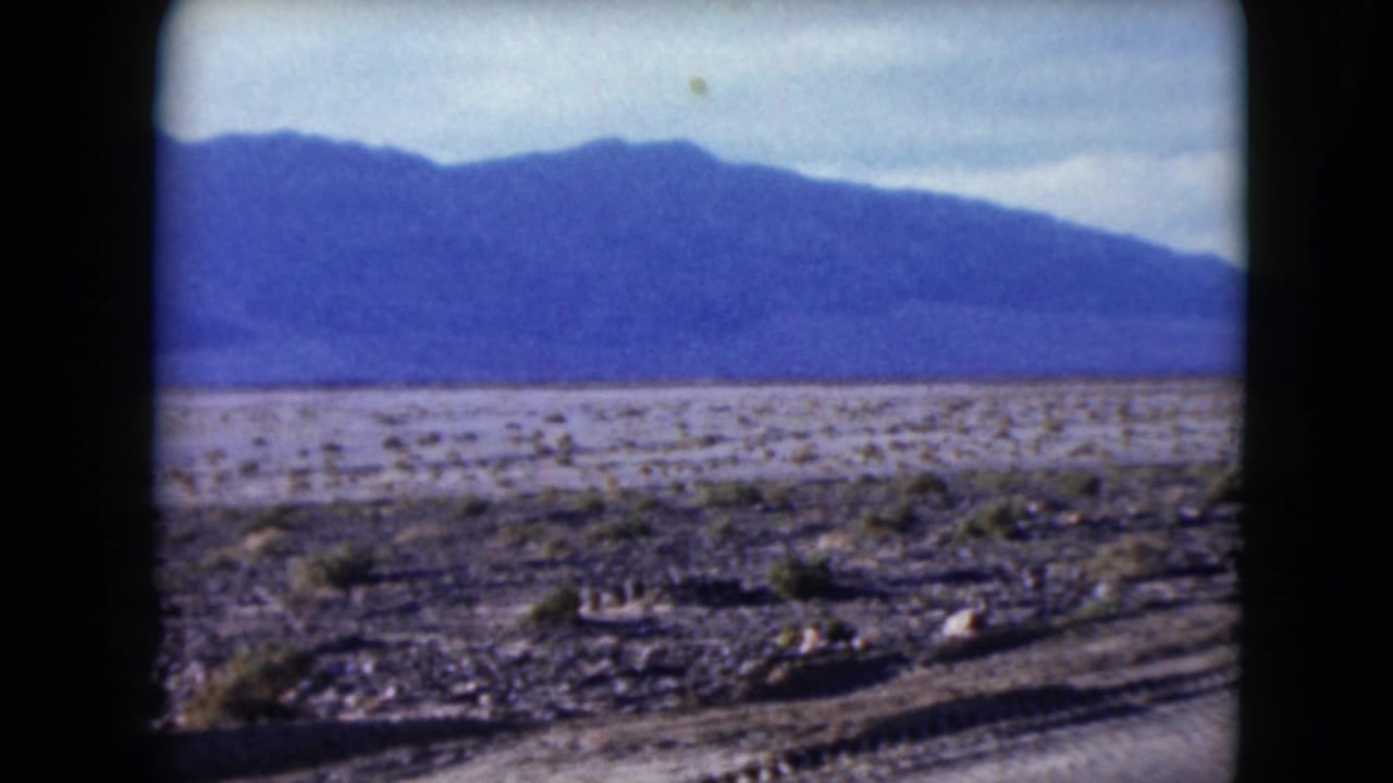 Desert scrubland and distant blue mountains seen beside a road through an 8mm frame.