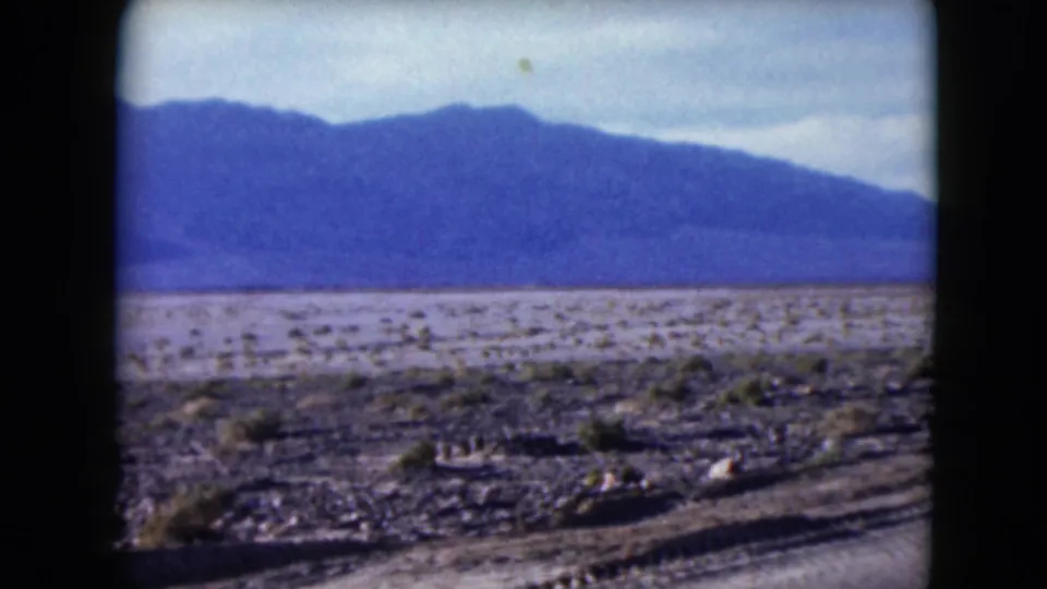 1961 Nevada 8mm view of sparse desert ground and a hazy mountain range beyond the roadside.