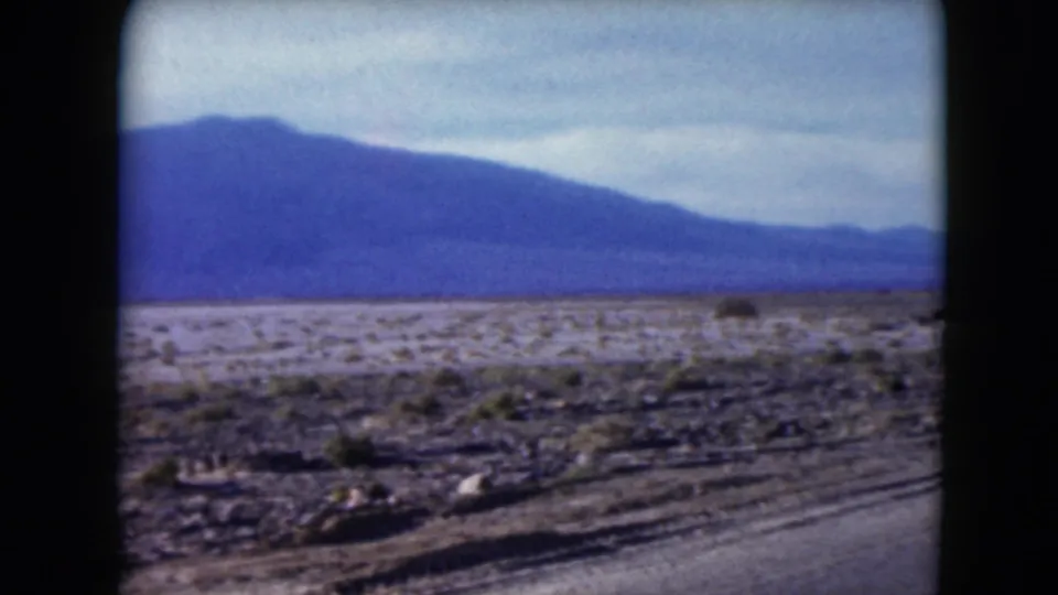 1961 Nevada home-movie frame showing open desert scrub and a hazy mountain range from a moving vehicle.