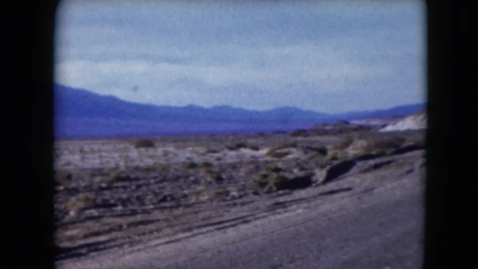 1961 Nevada travel view with a paved road edge, sparse desert vegetation, and distant mountains.