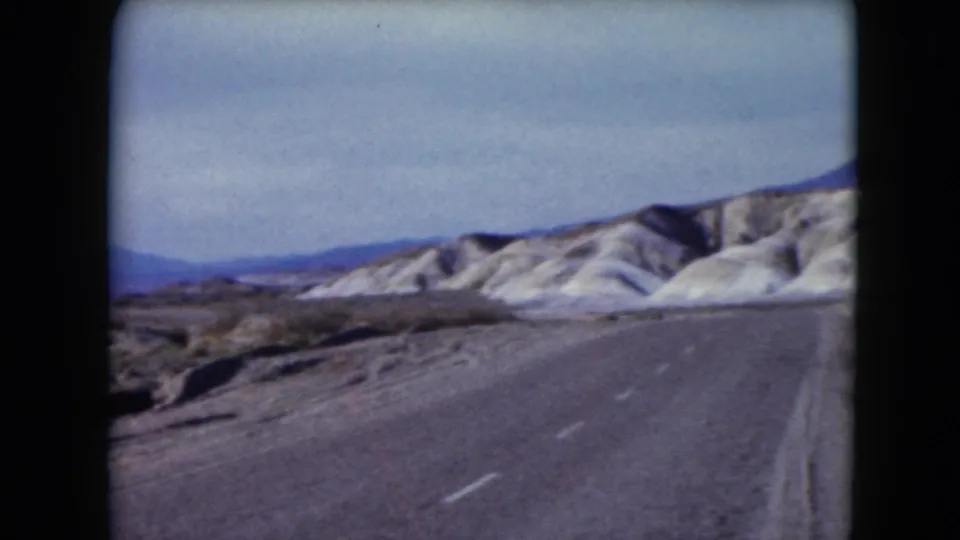 1961 Nevada 8mm frame of a desert road curving past light-colored hills with mountains in the background.