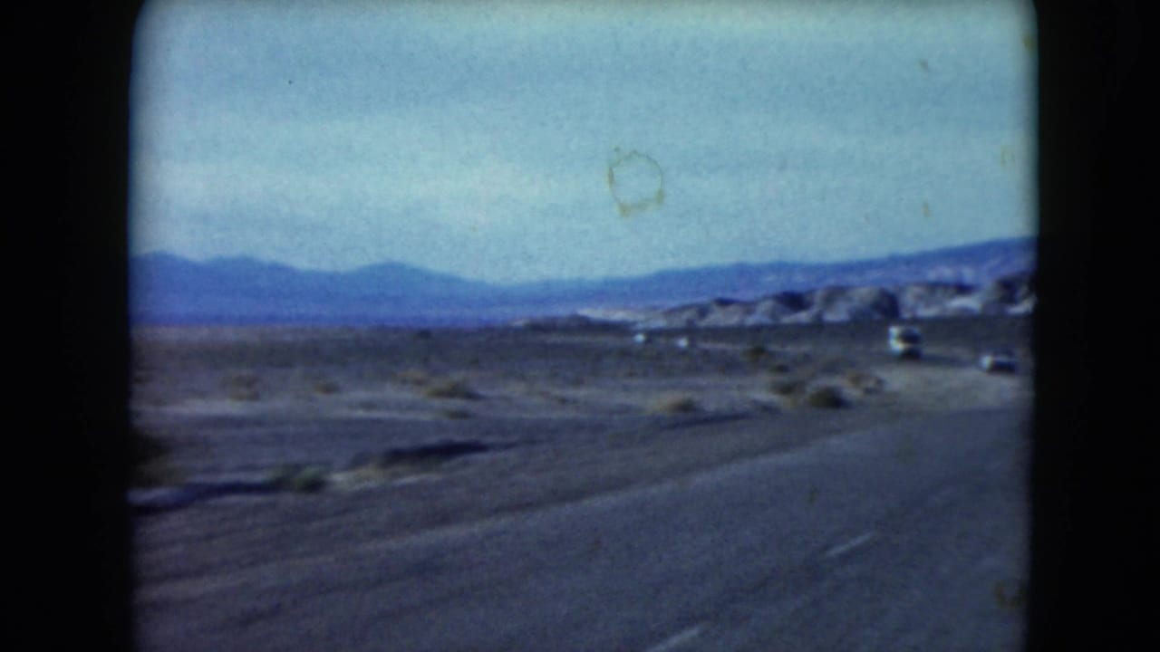 Paved desert road with distant cars, scrubland, mountains, and 8mm frame borders.