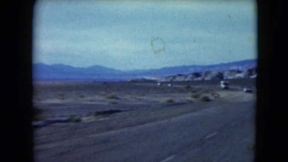 1961 Nevada 8mm road-trip frame showing a paved desert highway, distant cars, low shrubs, mountains, and faded blue sky.