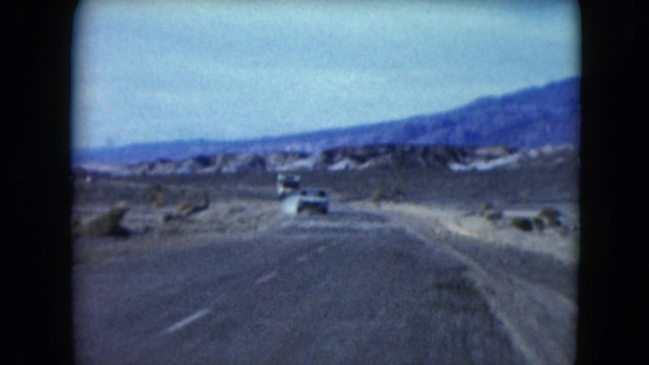 1961 Nevada desert highway view with cars traveling ahead, a dusty roadside, low vegetation, and mountains beyond.