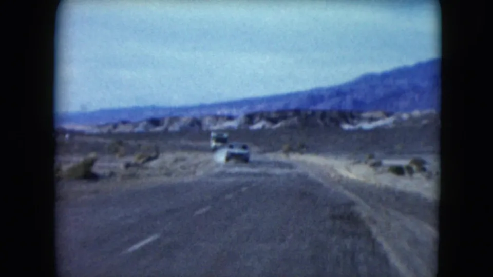 1961 Nevada desert highway view with cars traveling ahead, a dusty roadside, low vegetation, and mountains beyond.