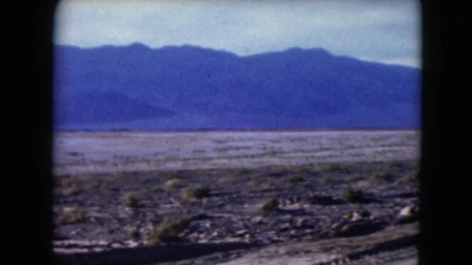 1961 Nevada desert landscape frame showing open arid ground, sparse shrubs, rocky foreground, and a broad mountain range.