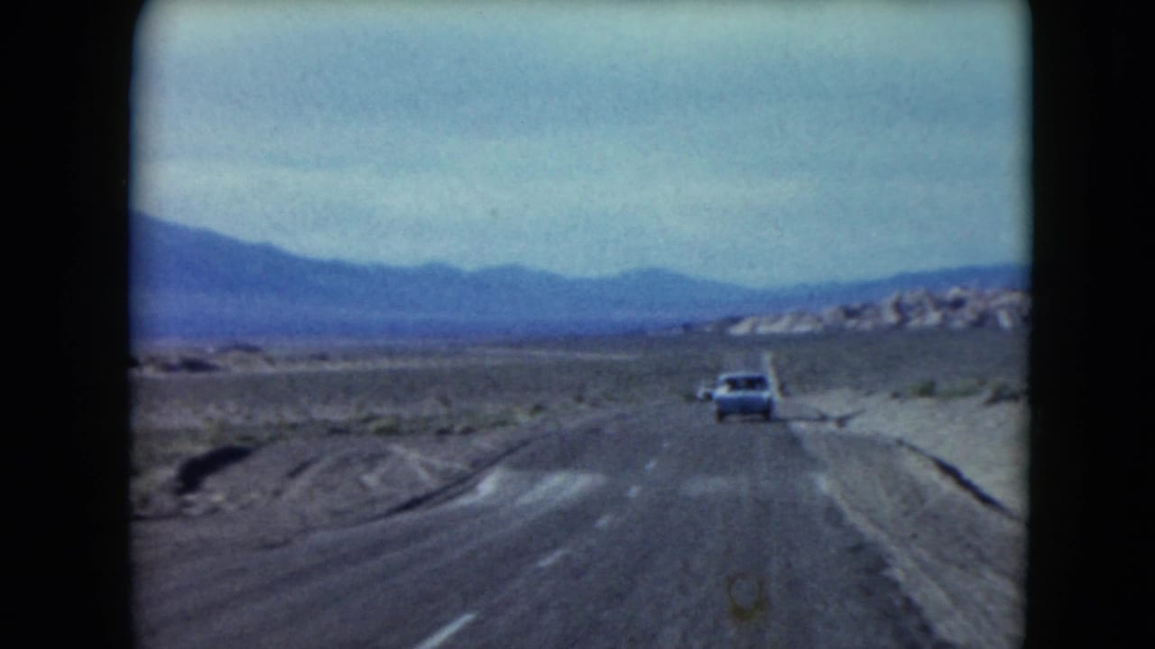 1961 Nevada 8mm frame showing a forward view along a desert highway, with a white car ahead, dry roadside terrain and distant mountains.