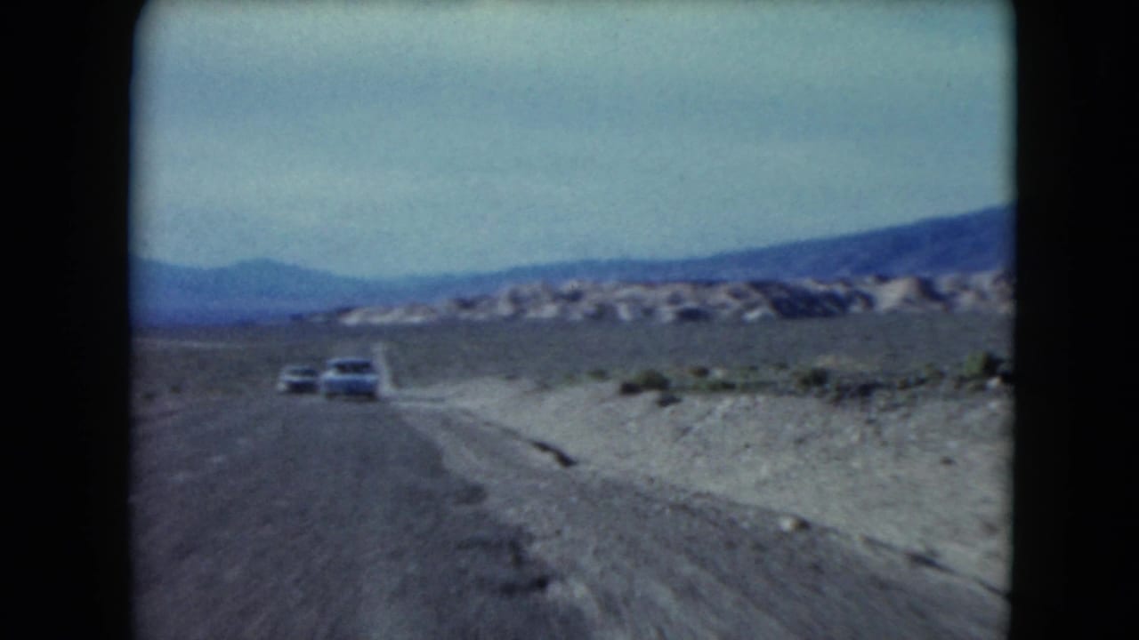 1961 Nevada road-trip frame with a white car on a two-lane highway, arid roadside ground, sparse shrubs and distant mountain ridges.