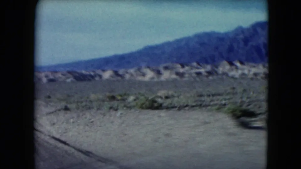 1961 Nevada 8mm frame from a moving vehicle, showing blurred roadside desert, sparse plants, low rocky hills and mountains under a pale sky.