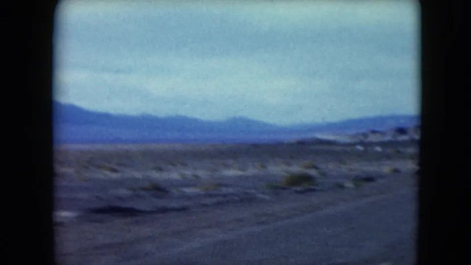 1961 Nevada desert landscape frame with the road edge in the foreground, sparse vegetation across open ground and distant blue mountain ranges.