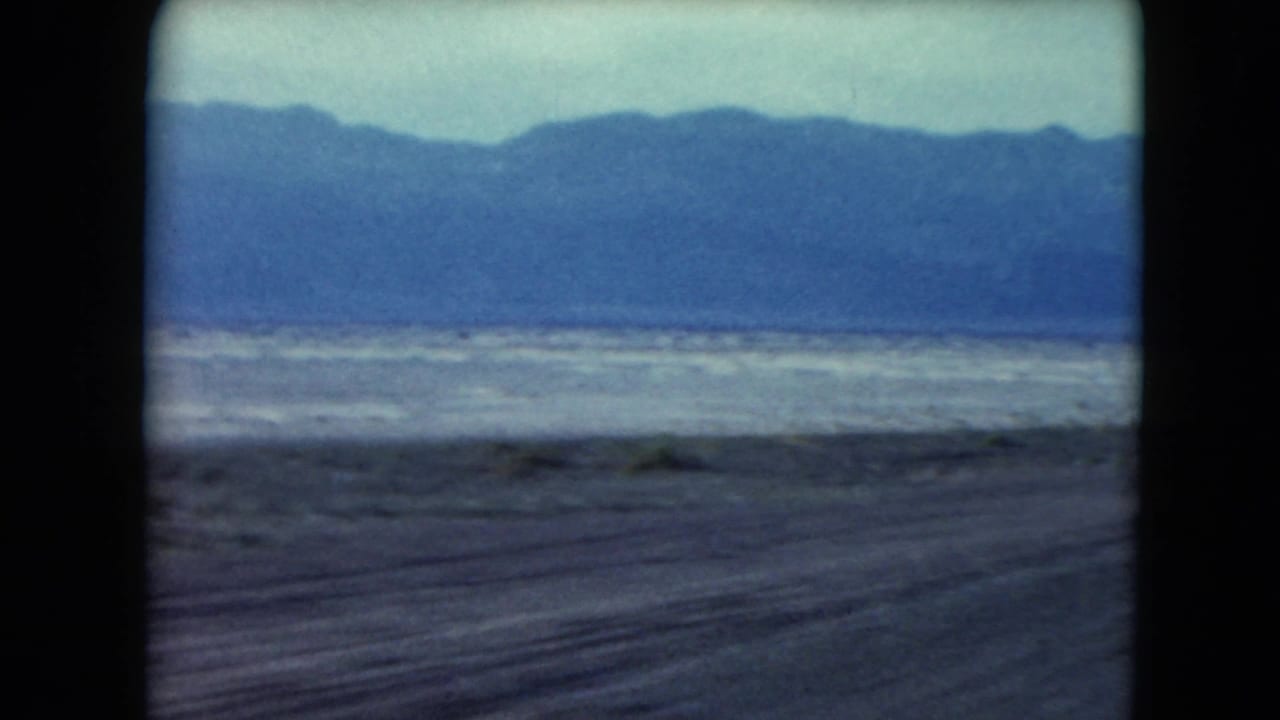 Desert highway seen from a moving vehicle with distant mountains and pale sky.