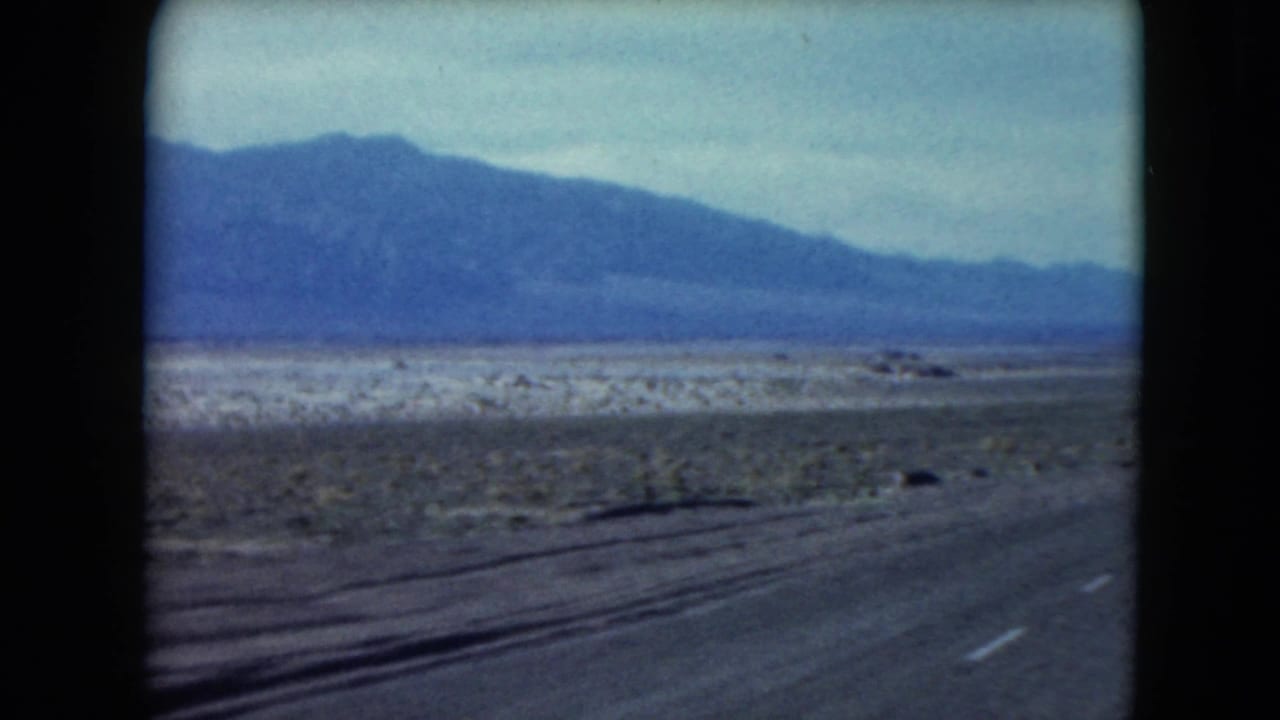 1961 Nevada desert highway view with dashed road markings, barren roadside terrain, and a mountain range beyond.