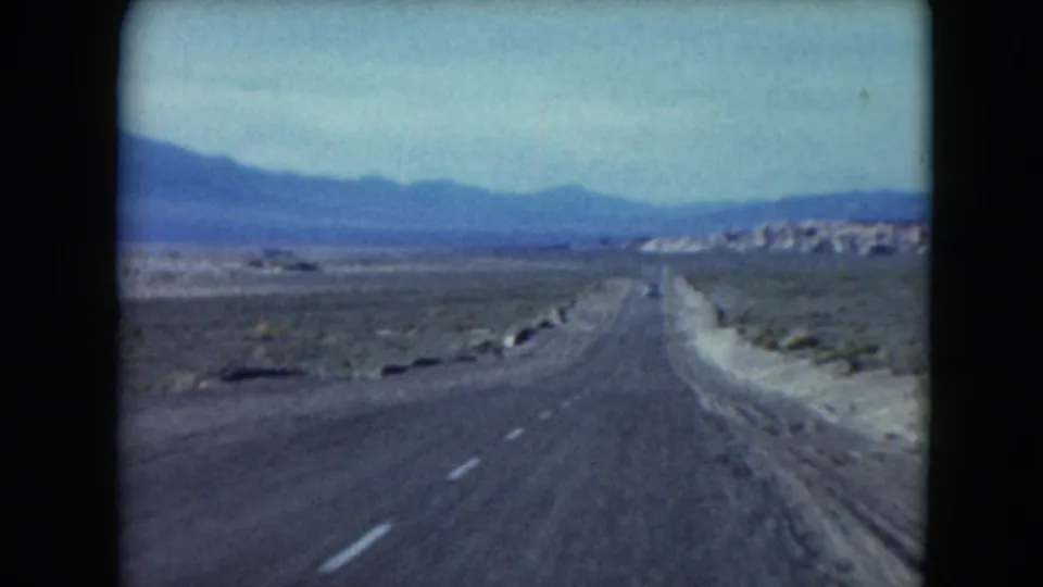 1961 Nevada home-movie frame of a straight desert highway leading toward distant structures and mountain ridges.
